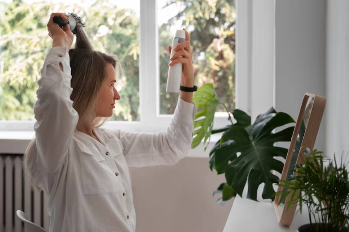 A person in a white shirt is styling their hair with a brush while holding a hair product, surrounded by indoor plants and bright windows.