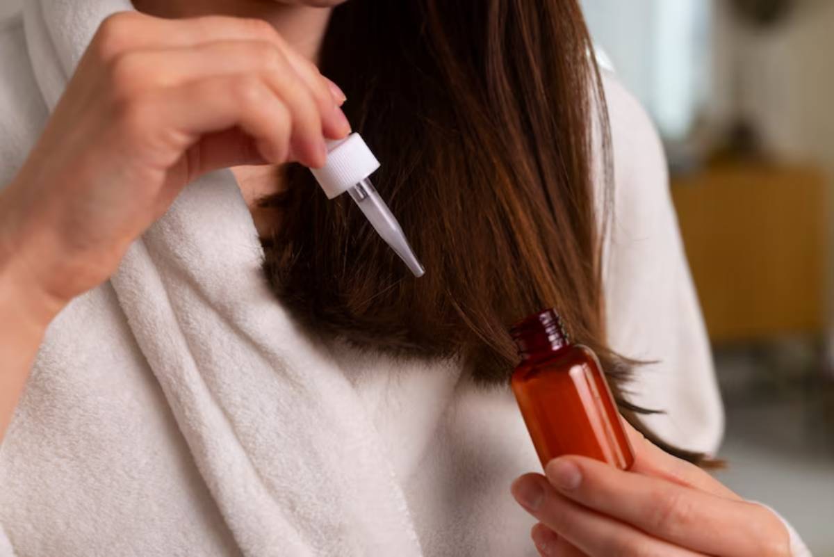 A woman in a white robe applies hair oil from a dropper into her dark, straight hair, focusing on a strand.