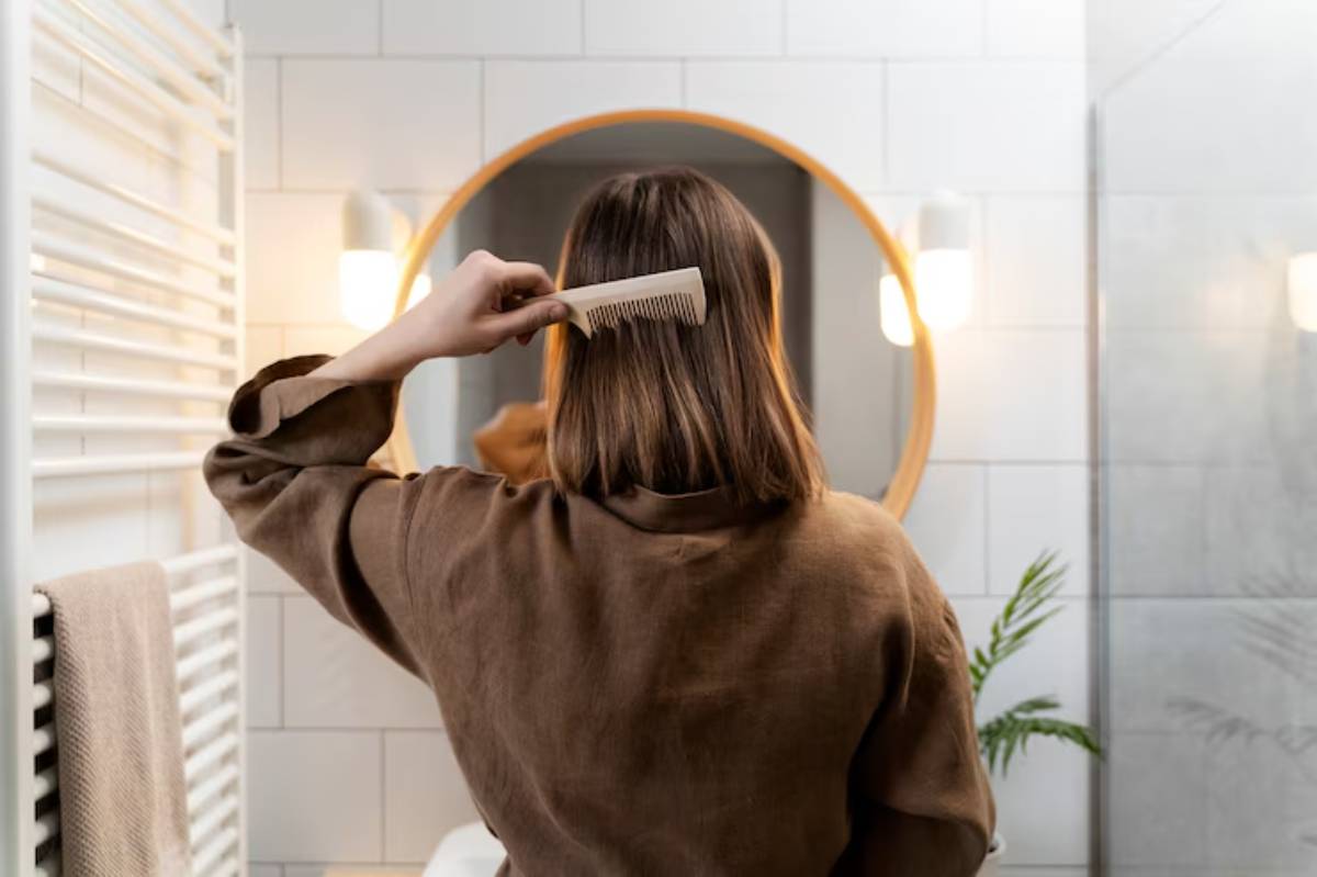 A woman with shoulder-length hair combs her hair in a modern bathroom, reflected in a circular mirror.