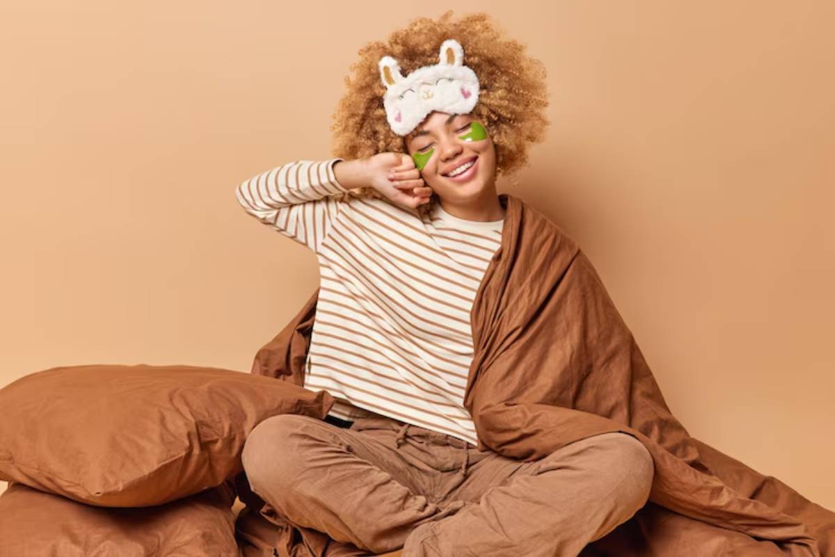 A person sits comfortably among large brown pillows, wearing a striped shirt and a cozy blanket, against a warm beige background.