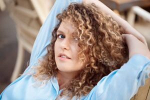 A woman with curly hair relaxes on a chair, dressed in a light blue shirt, enjoying a leisurely moment outdoors.