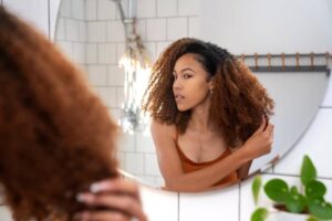 A person with long, curly hair is grooming themselves in front of a mirror in a bright bathroom with green plants nearby.