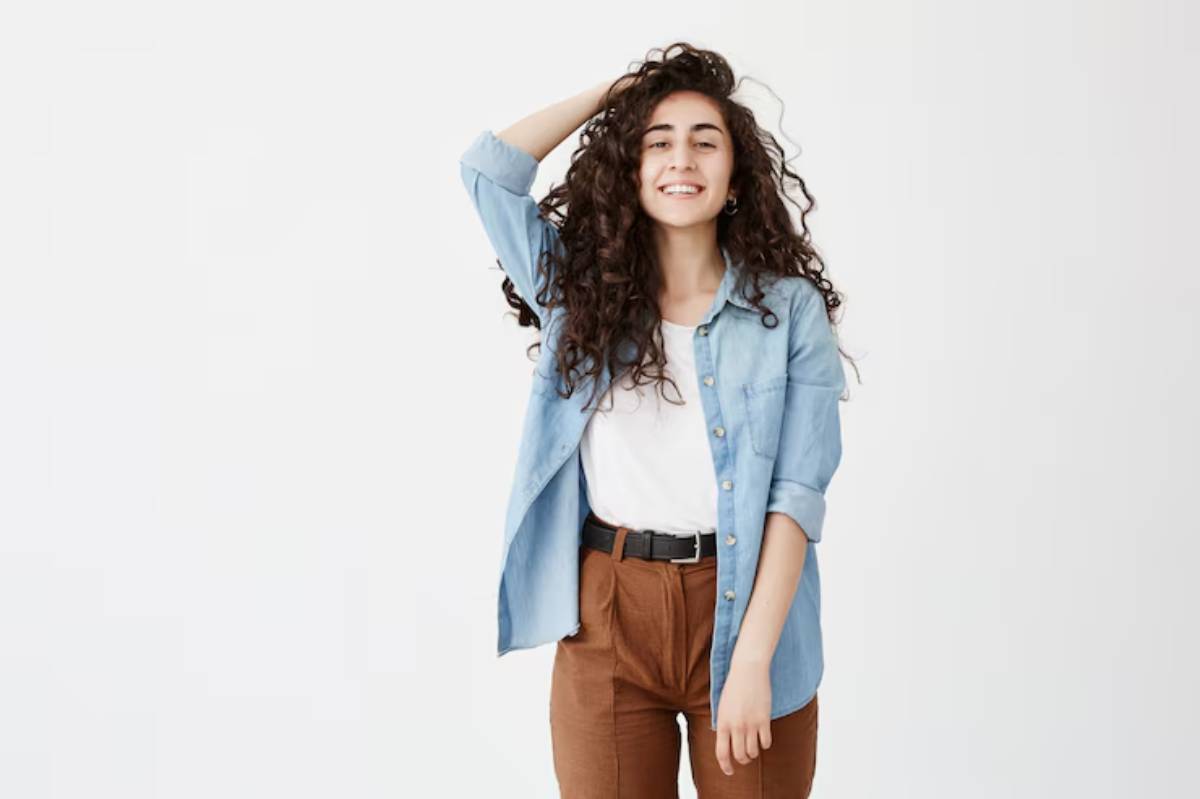 A person with curly hair poses in a light blue button-down shirt and brown high-waisted pants against a plain white background.