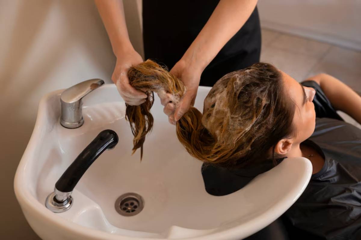 A stylist washes a client's long hair with shampoo at a salon, with bubbly suds in a hair washing station.