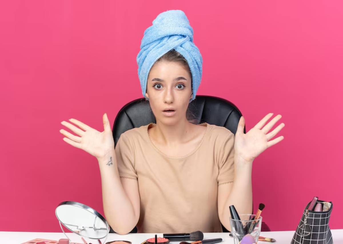 A woman in a beige shirt and blue towel wraps raises her hands in a bright pink room filled with cosmetics and a mirror.