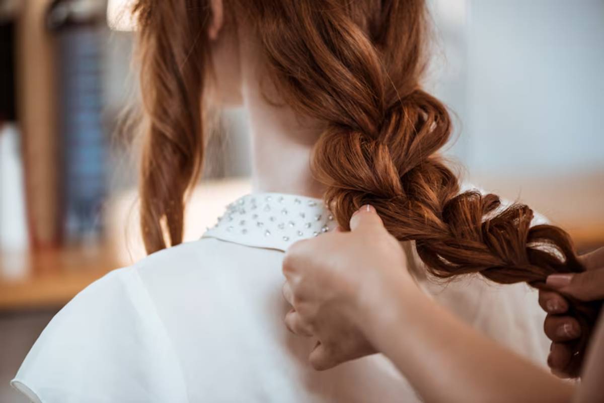 A close-up of a person braiding the long, red hair of a girl wearing a white blouse with embellishments.