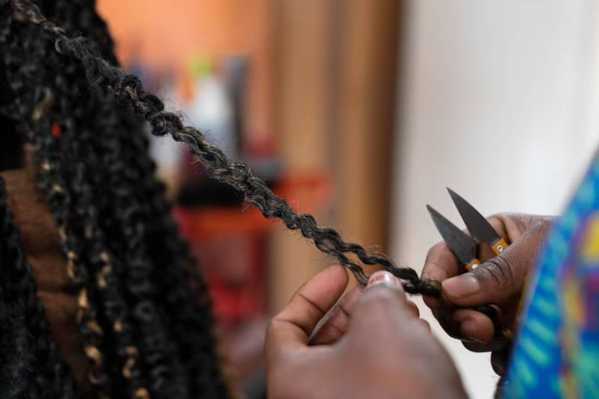 A close-up of hands holding a braided hair strand, with scissors positioned to cut the braid, set in a vibrant salon environment.