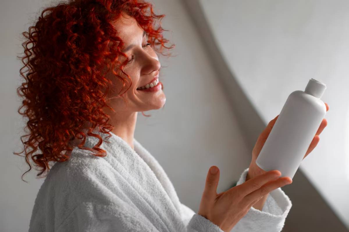 A person in a white bathrobe holds a plain white bottle, gazing thoughtfully with curly red hair in a softly lit setting.