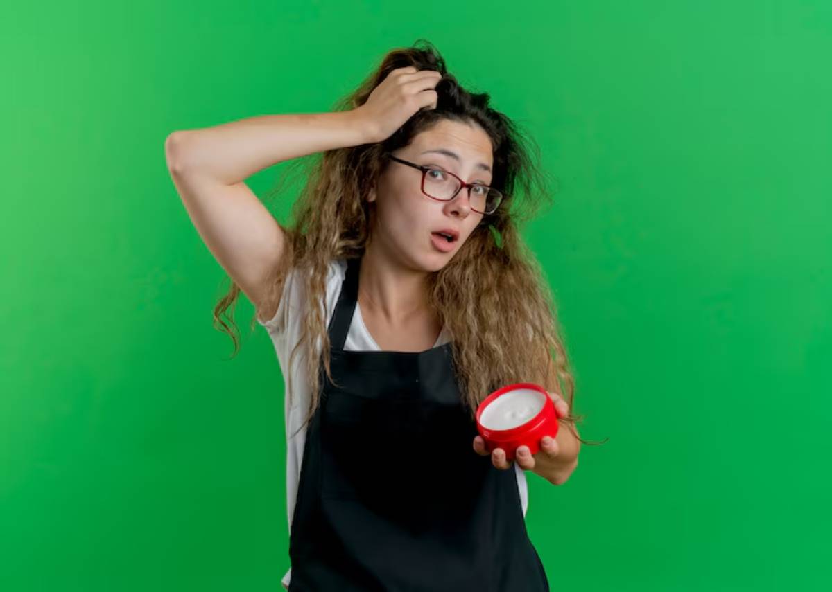 A person with curly hair holds a red container while their other hand rests on their head, set against a vibrant green background.