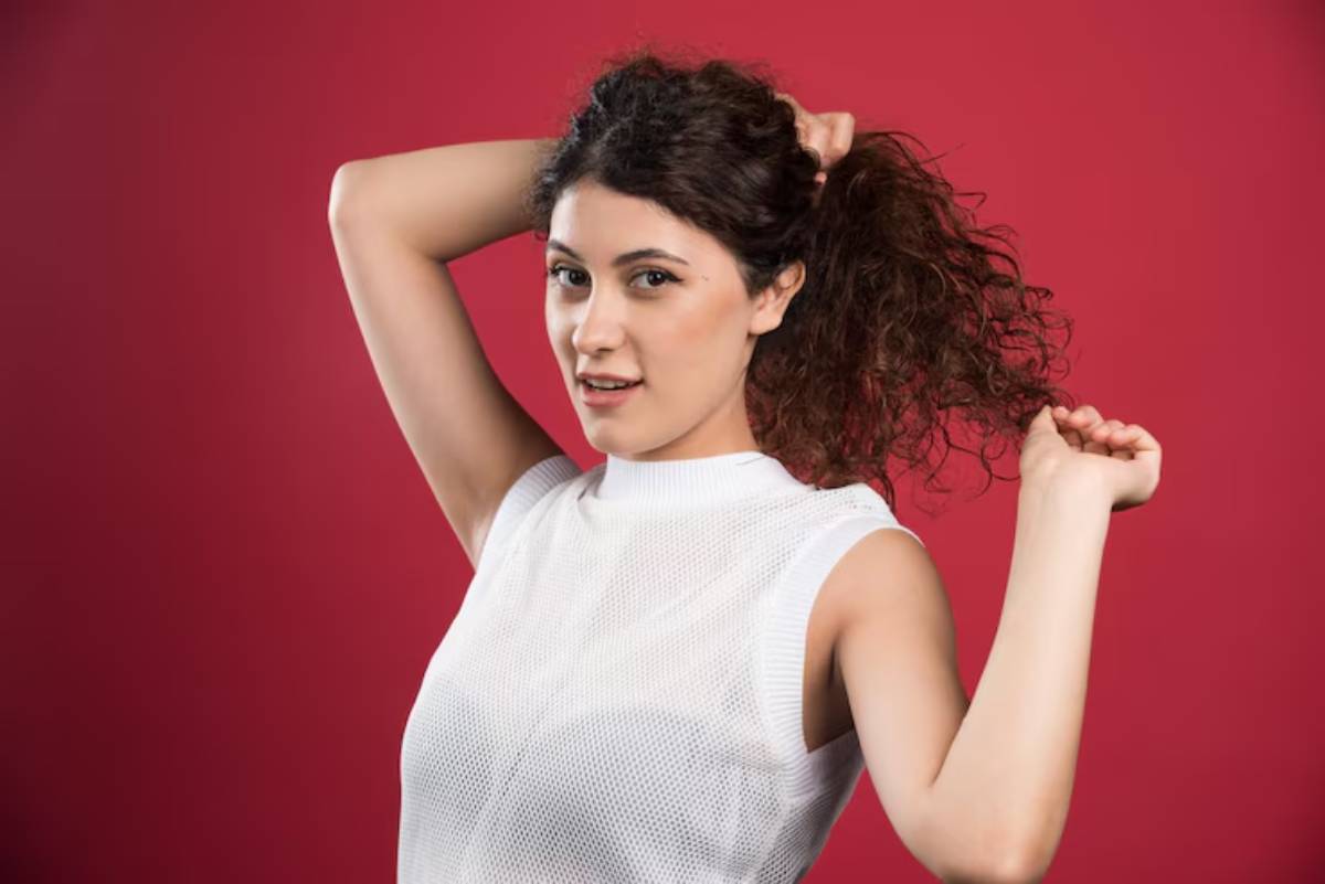 A young woman in a white sleeveless top plays with her curly hair against a vibrant red background.