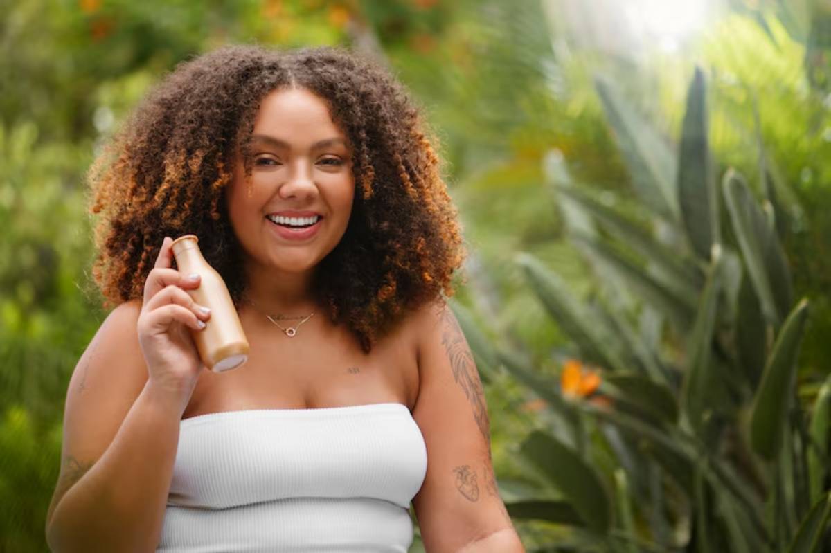 A woman with curly hair holds a gold bottle, surrounded by lush greenery and vibrant tropical plants. She wears a white strapless top.