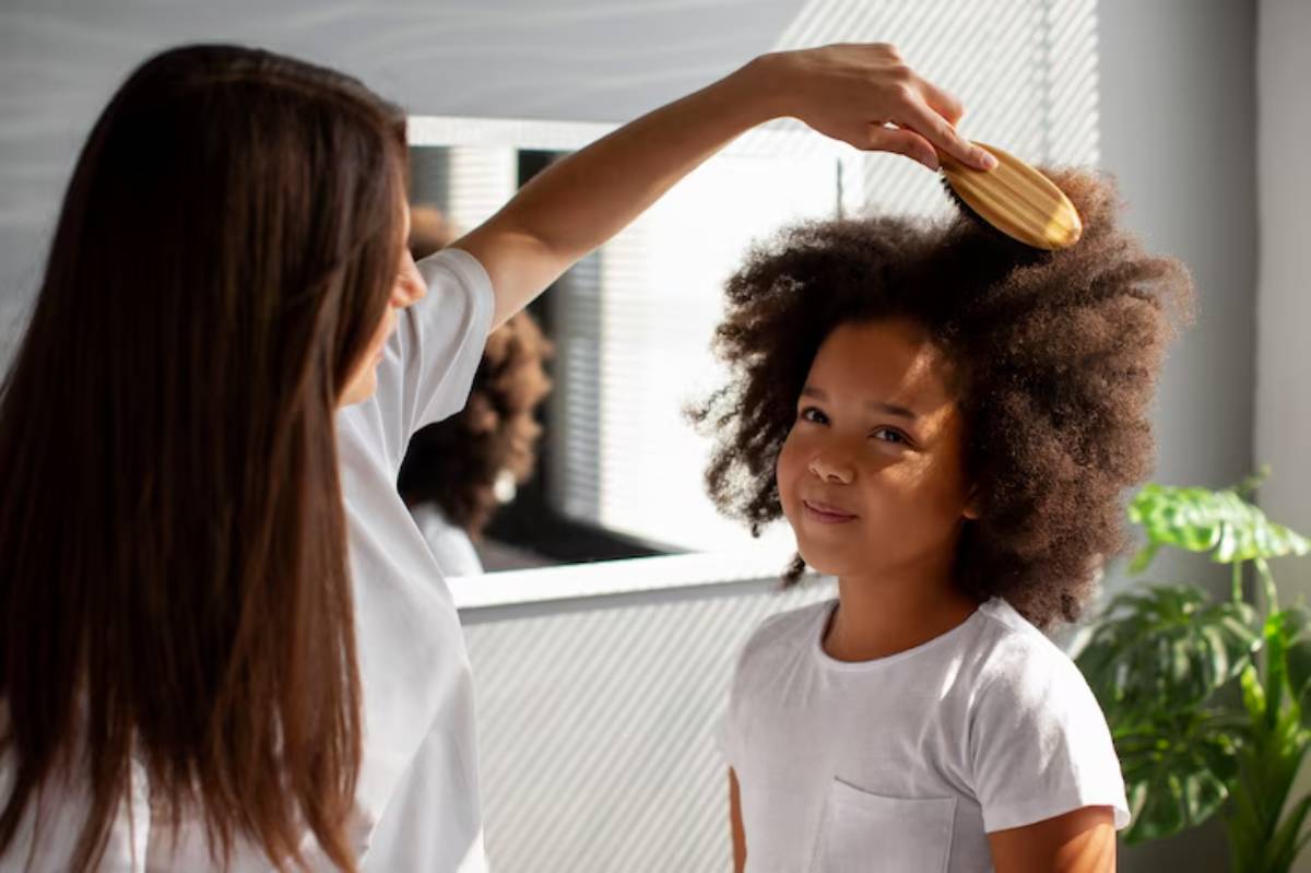 A person brushes a child's curly hair in a brightly lit room with a leafy plant and a large mirror in the background.