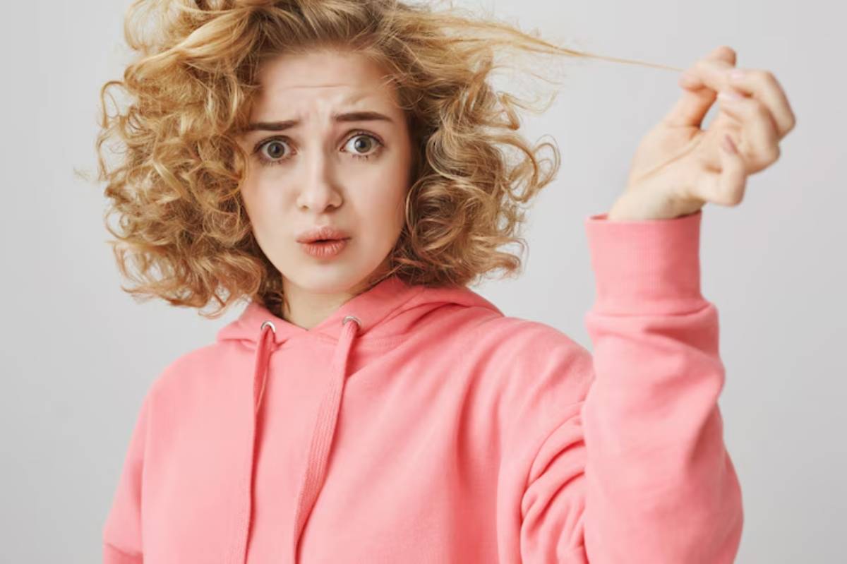 A person wearing a pink hoodie playfully twirls a strand of curly hair against a neutral background.