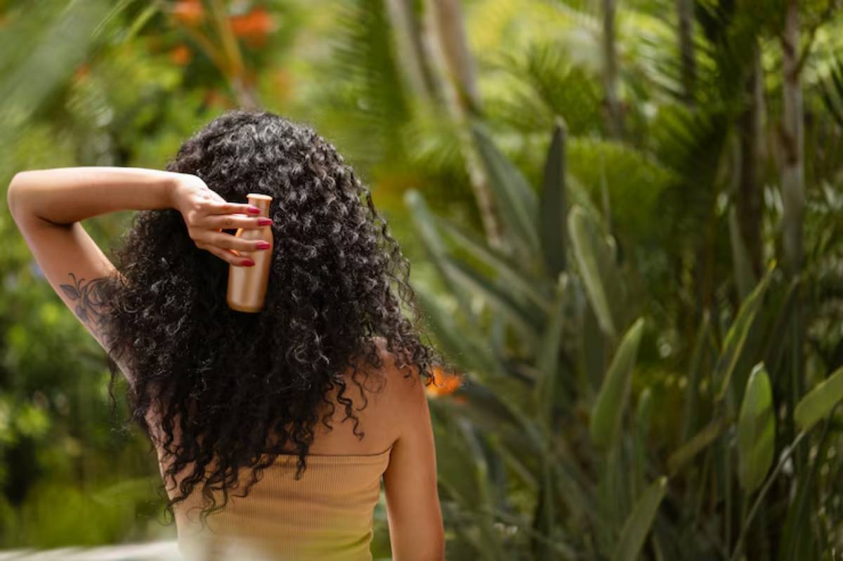 A woman with curly hair holds a golden bottle, surrounded by lush greenery, enjoying a moment in nature.