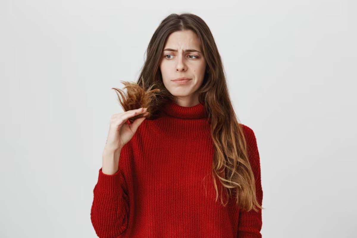 A woman in a red sweater holds a section of her long, wavy hair while posing against a plain white background.