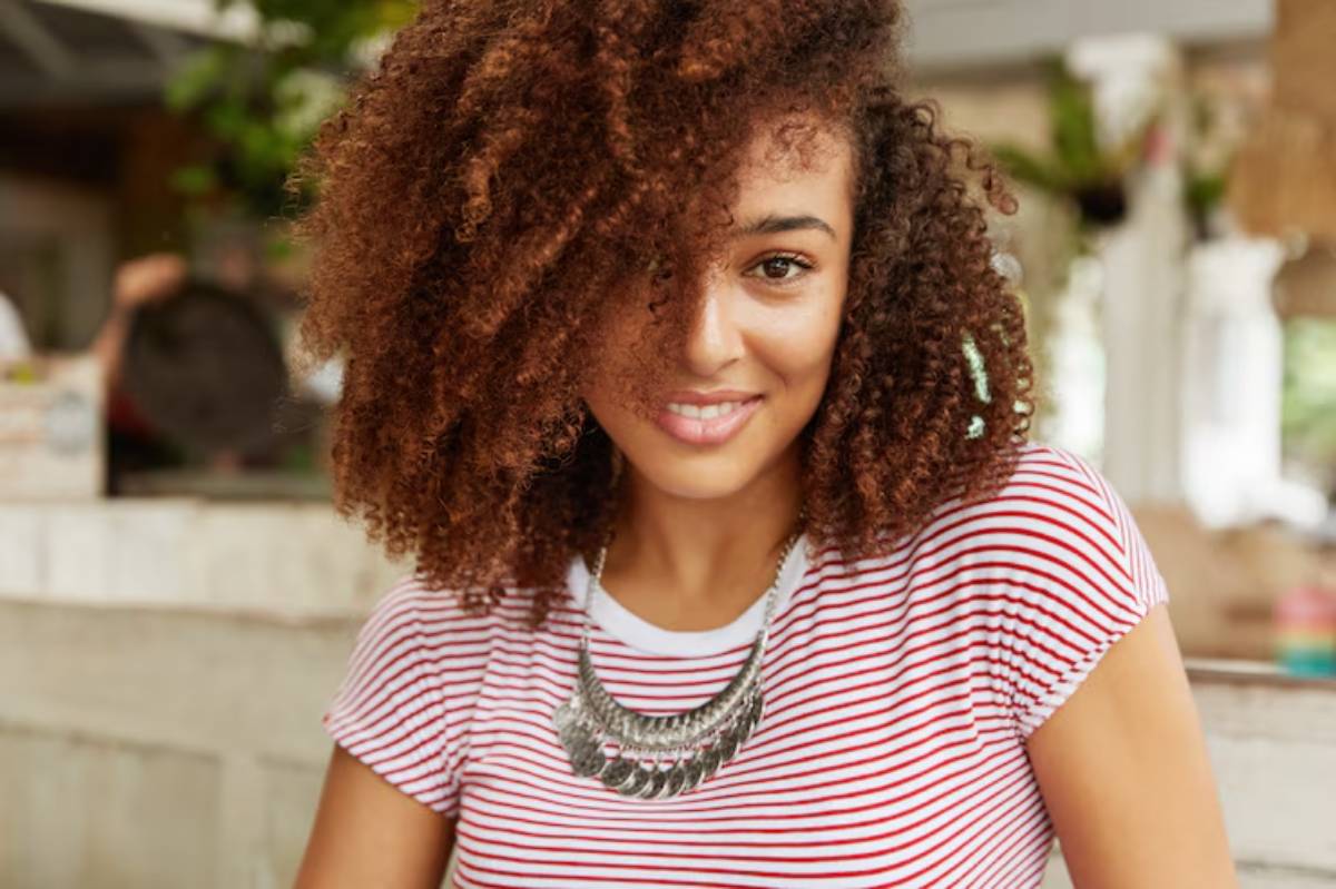 A person with curly, brown hair wearing a striped shirt and a statement necklace, sitting in a cozy café setting. 