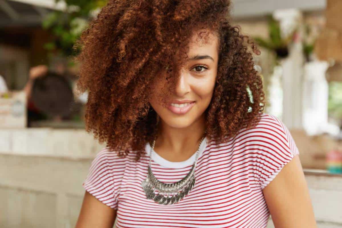 A person with curly, brown hair wearing a striped shirt and a statement necklace, sitting in a cozy café setting.
