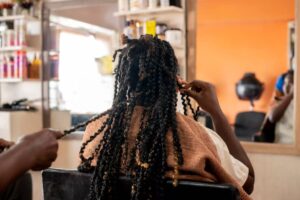 A person with long braided hair sits in a salon, while a stylist expertly works on their hair, surrounded by colorful hair products.