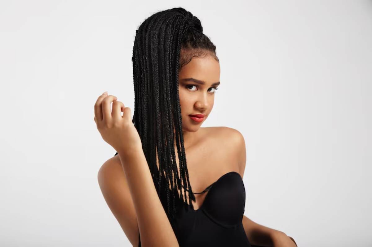 A young woman with long box braids poses in a black top against a clean white background, showcasing her elegant hand gesture.