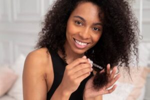 A person with curly hair holds a dropper, applying product to their hair while sitting in a softly lit, cozy room.