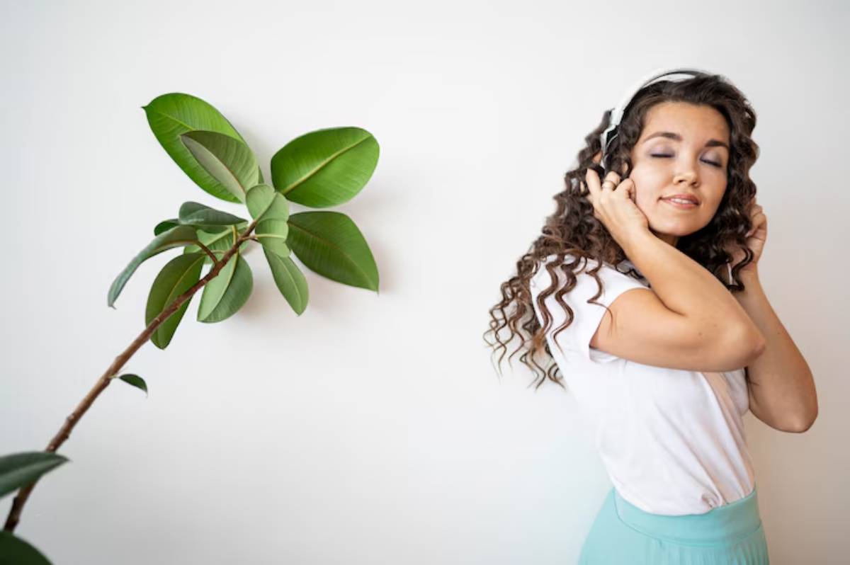 A woman with curly hair wears headphones, standing beside a large green plant against a white wall. 