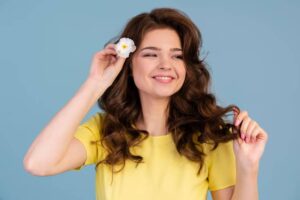 A woman with wavy brown hair, wearing a yellow shirt, holds a flower near her head against a light blue background.