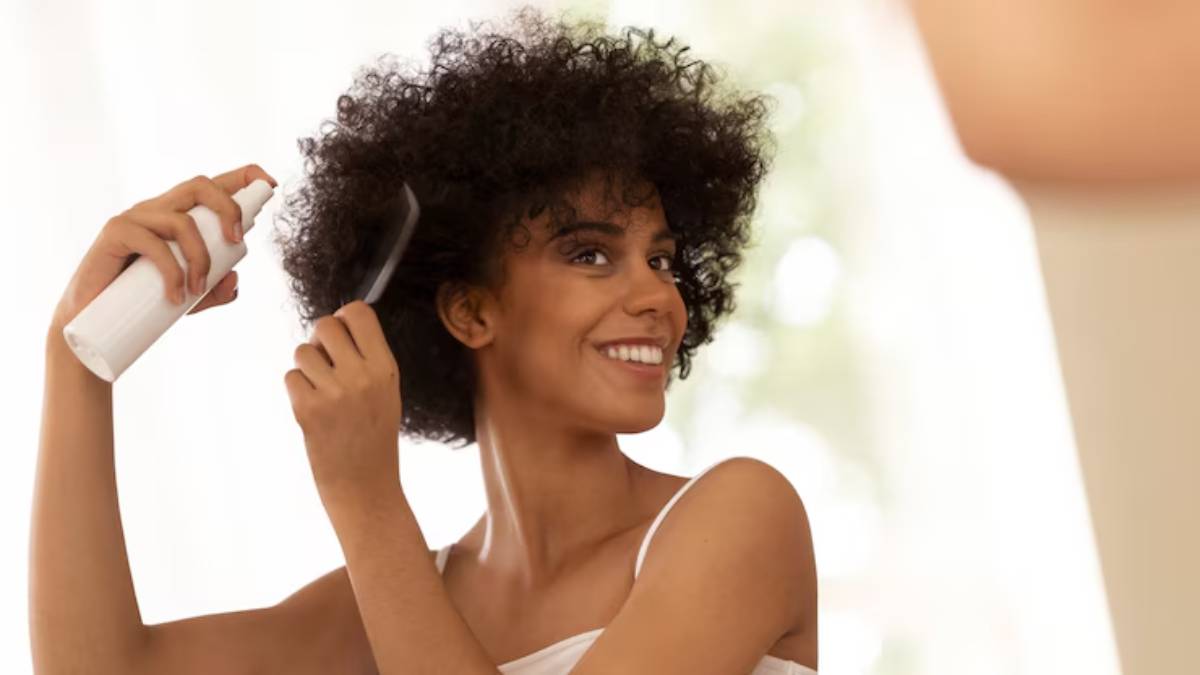 A person with curly hair uses a comb and a spray bottle while styling their hair in a bright, well-lit room.