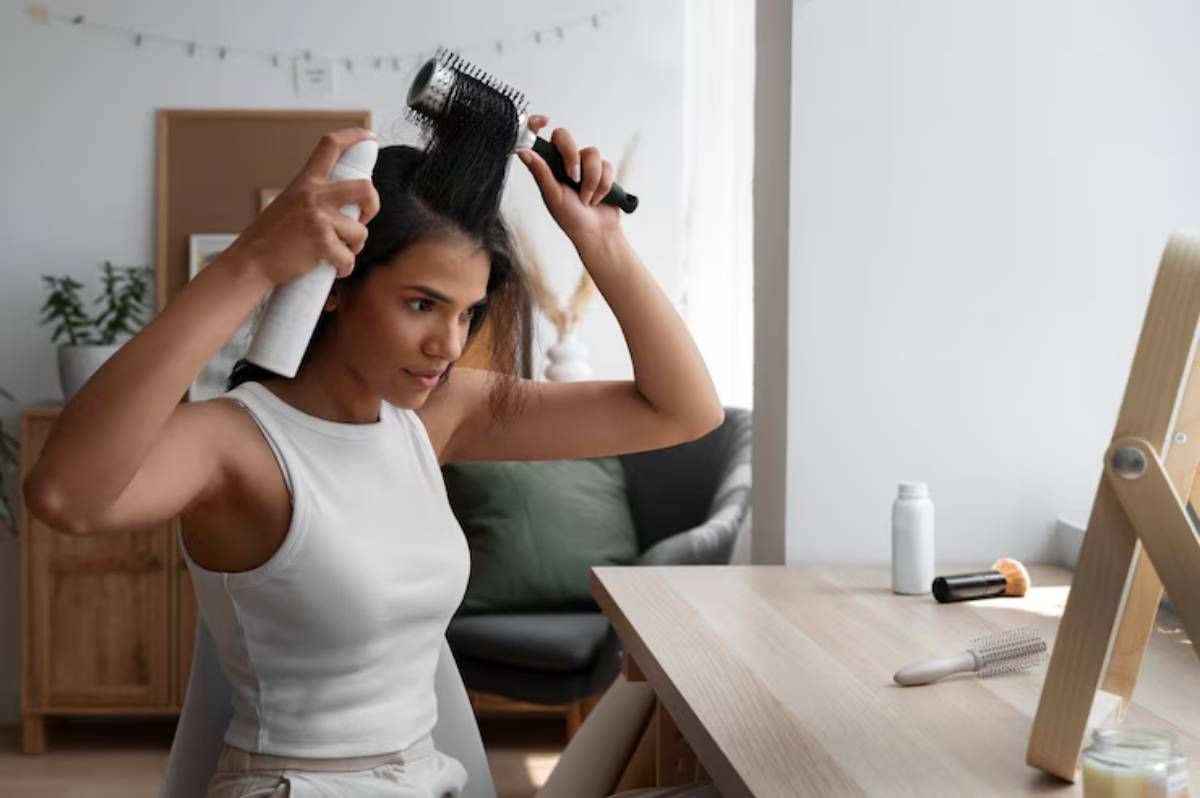 A person applies hair spray while styling their hair with a round brush, seated at a bright vanity in a cozy, modern room.