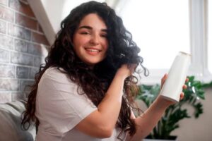 A person with curly hair holds a white bottle in one hand while adjusting their hair, sitting on a couch with houseplants nearby.