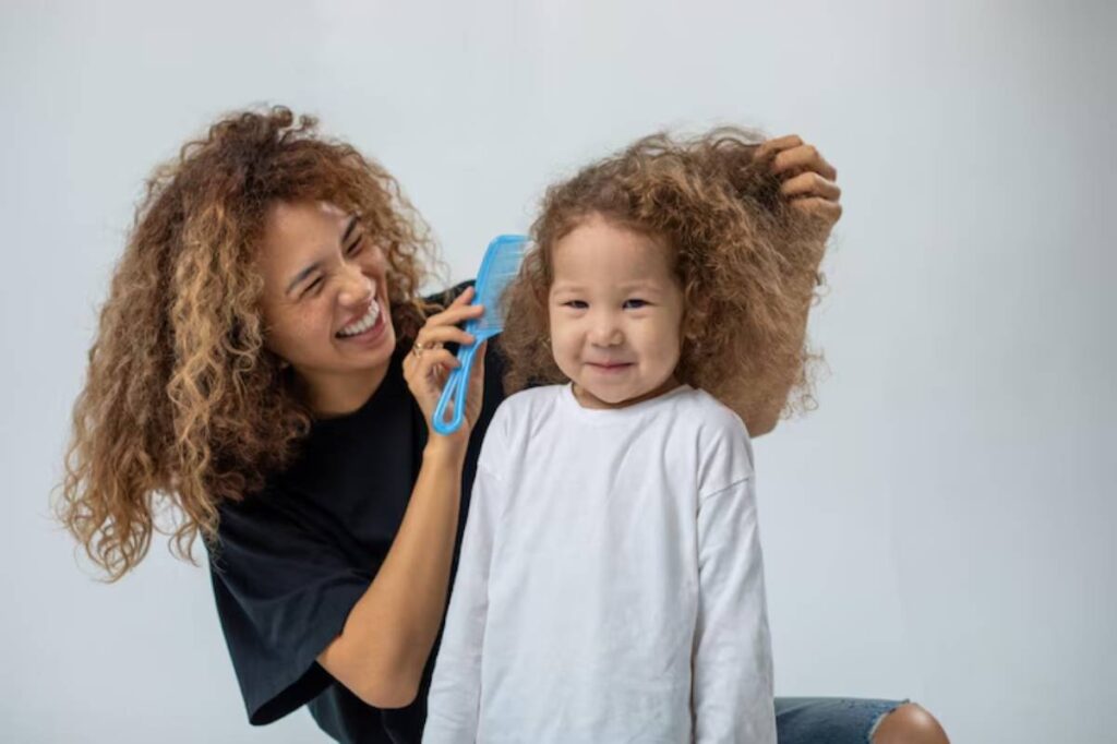 A woman with curly hair combs the hair of a child wearing a white shirt, both seated against a neutral background.