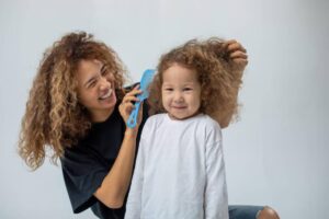 A woman with curly hair combs the hair of a child wearing a white shirt, both seated against a neutral background.