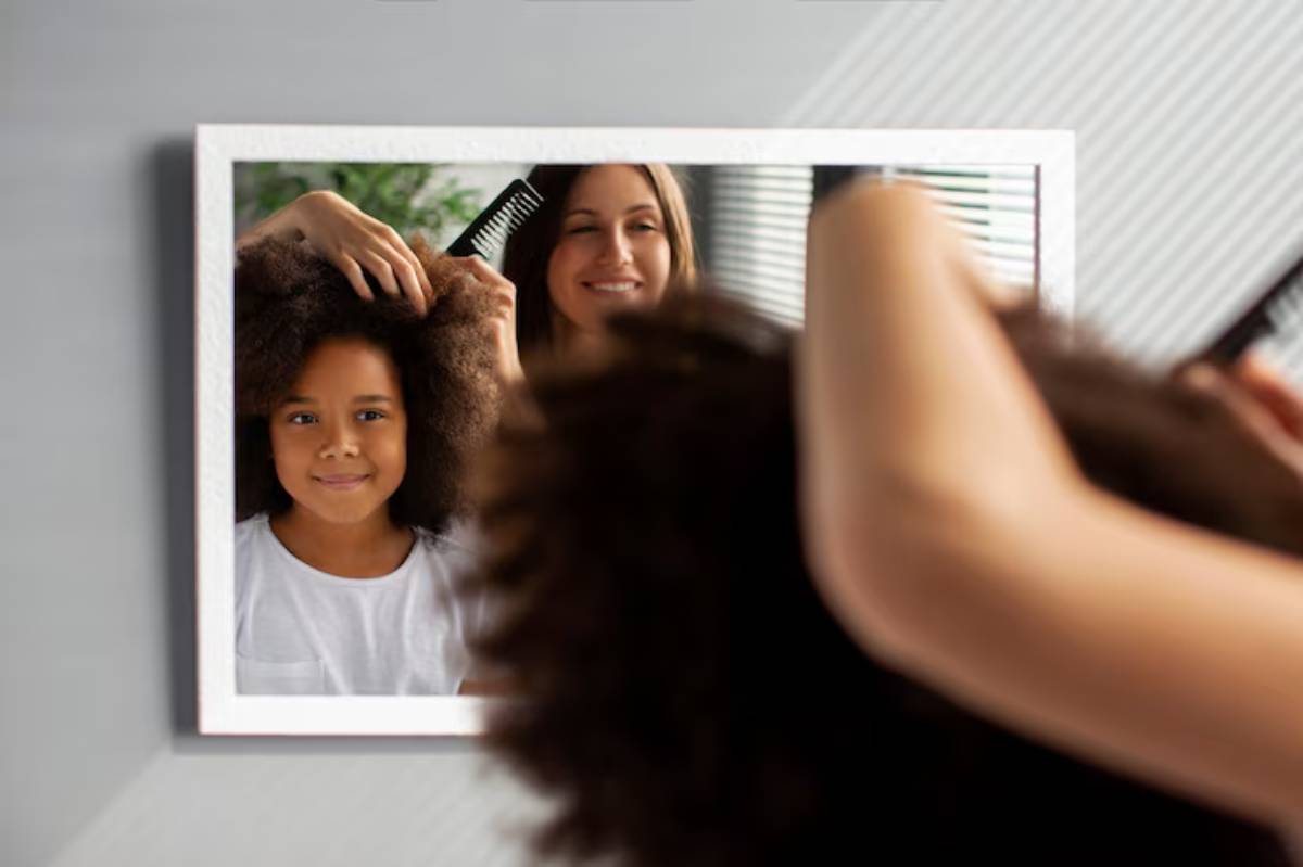 A person combs curly hair in front of a mirror, with soft natural light illuminating the scene and a minimalist background. 