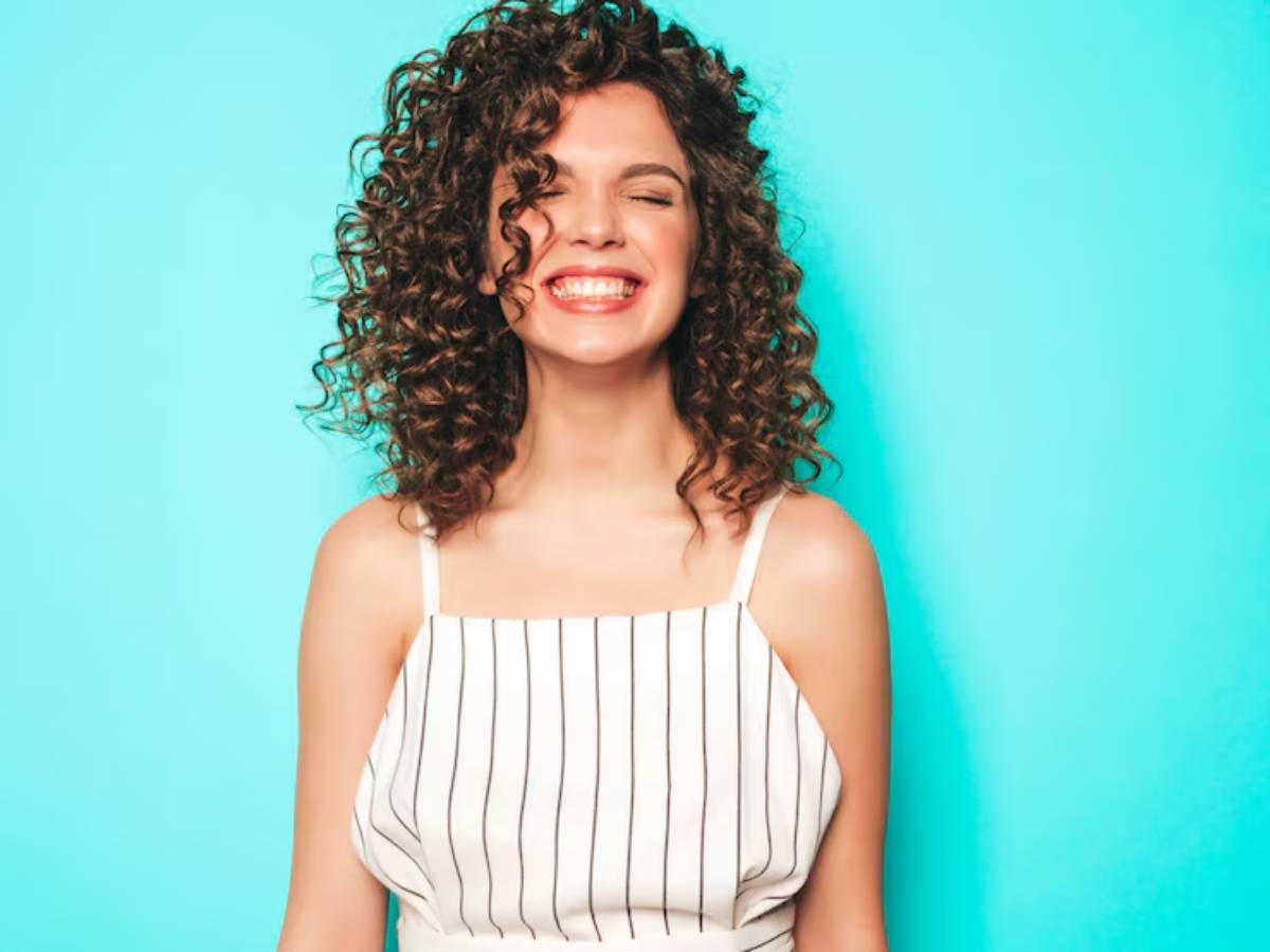  A woman with curly hair stands against a teal background, wearing a white striped tank top, showcasing a confident stance.