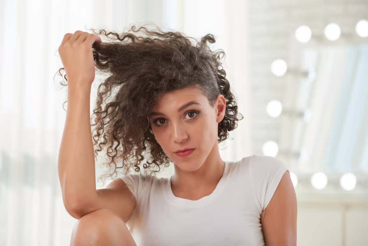 A person with curly hair holds their hair up, seated comfortably in a bright room with soft lighting and a simple backdrop.