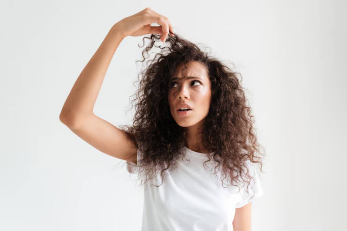 A person with curly hair holds a section of their hair, wearing a plain white t-shirt against a minimalistic light background.