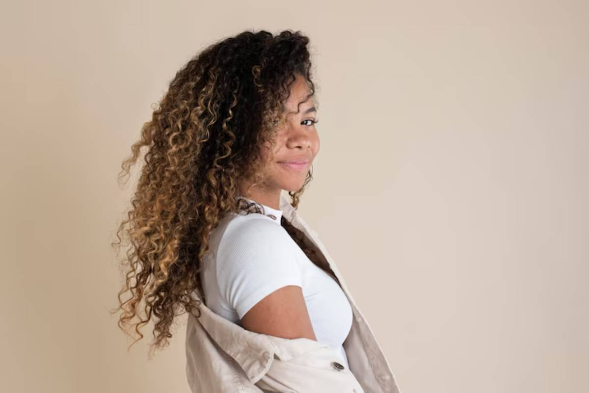 A woman with curly hair is wearing a white top and a beige jacket, facing sideways against a soft beige background.