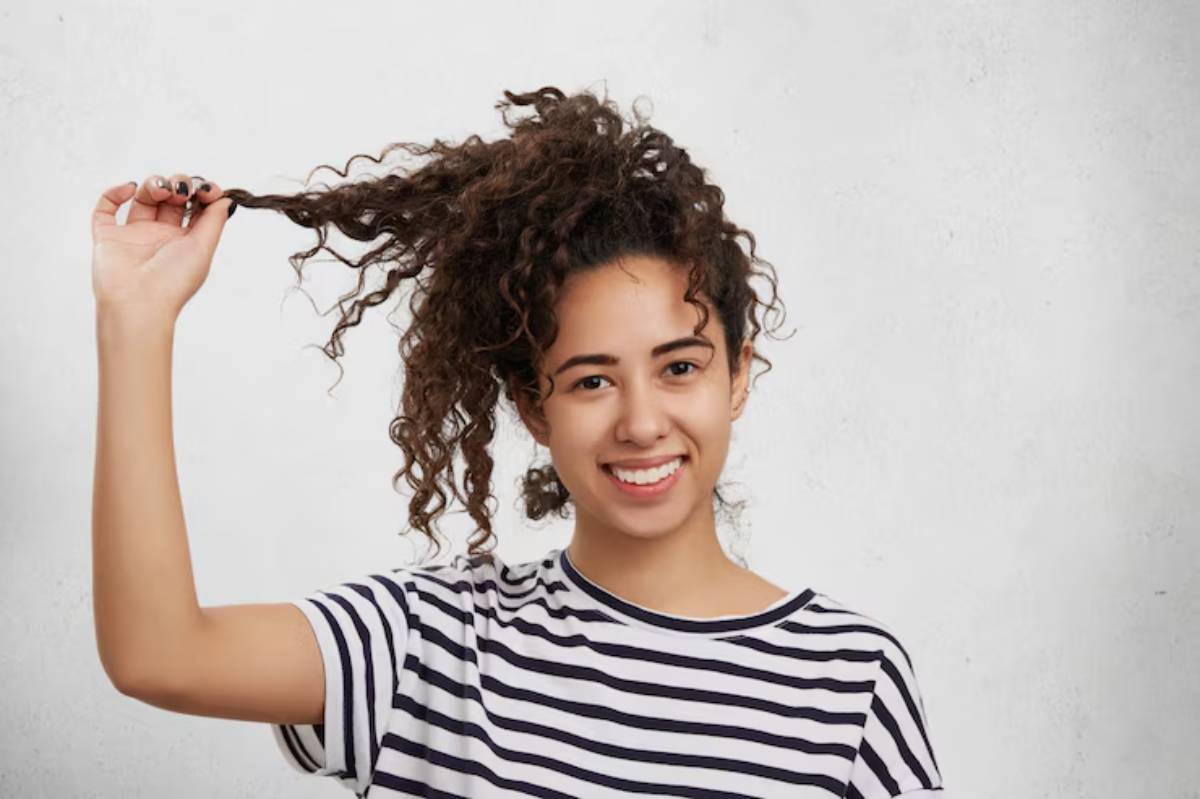 A person with curly hair in a striped shirt playfully holds up a section of hair against a neutral background.