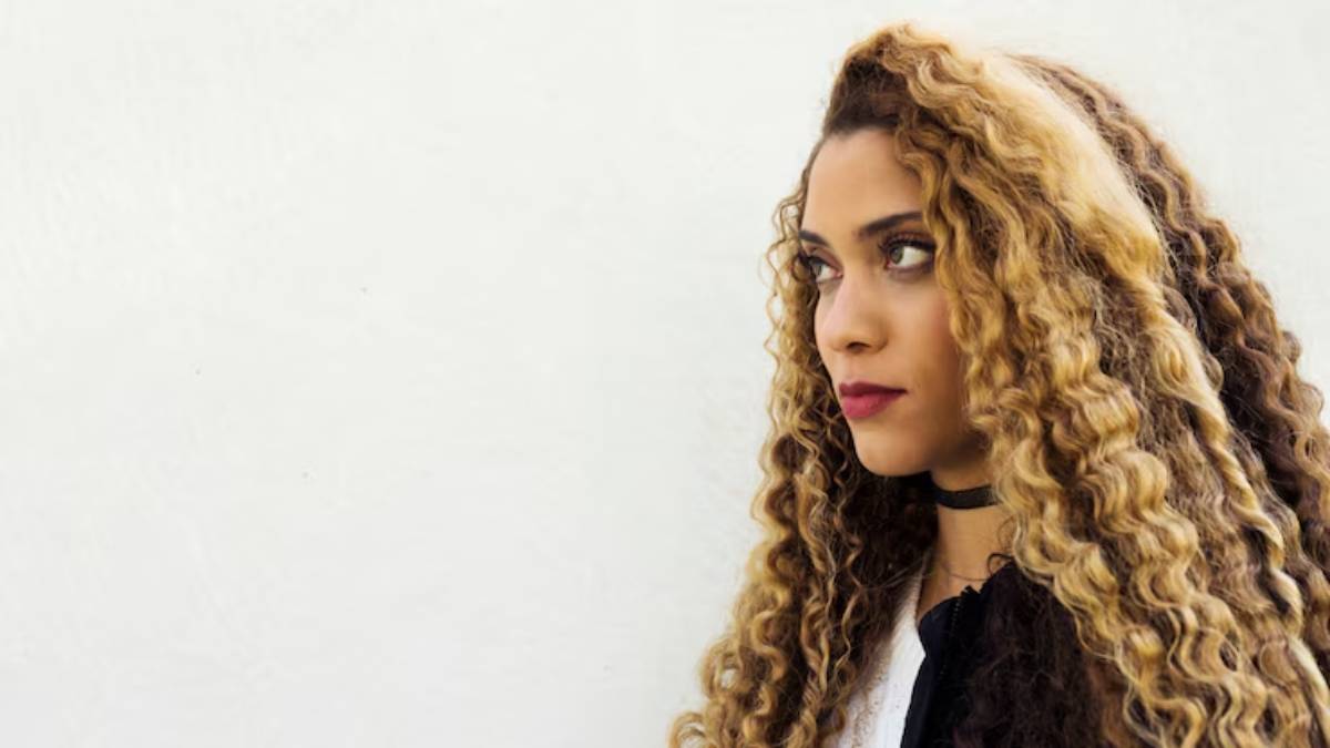 A person with long, voluminous curly hair stands against a plain white wall, wearing a black choker and casual attire. 