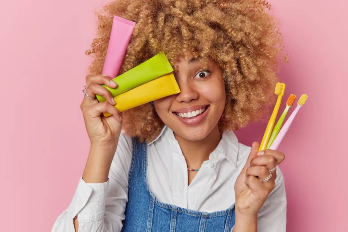 A person with curly hair holds colorful toothpaste tubes and vibrant toothbrushes against a pink background, promoting dental care. 