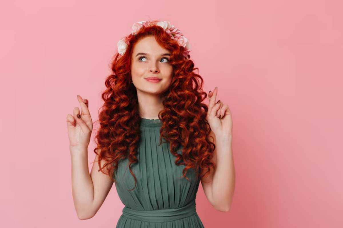 A woman with vibrant red, curly hair wearing a pleated green dress and a floral crown, posing against a pink background.