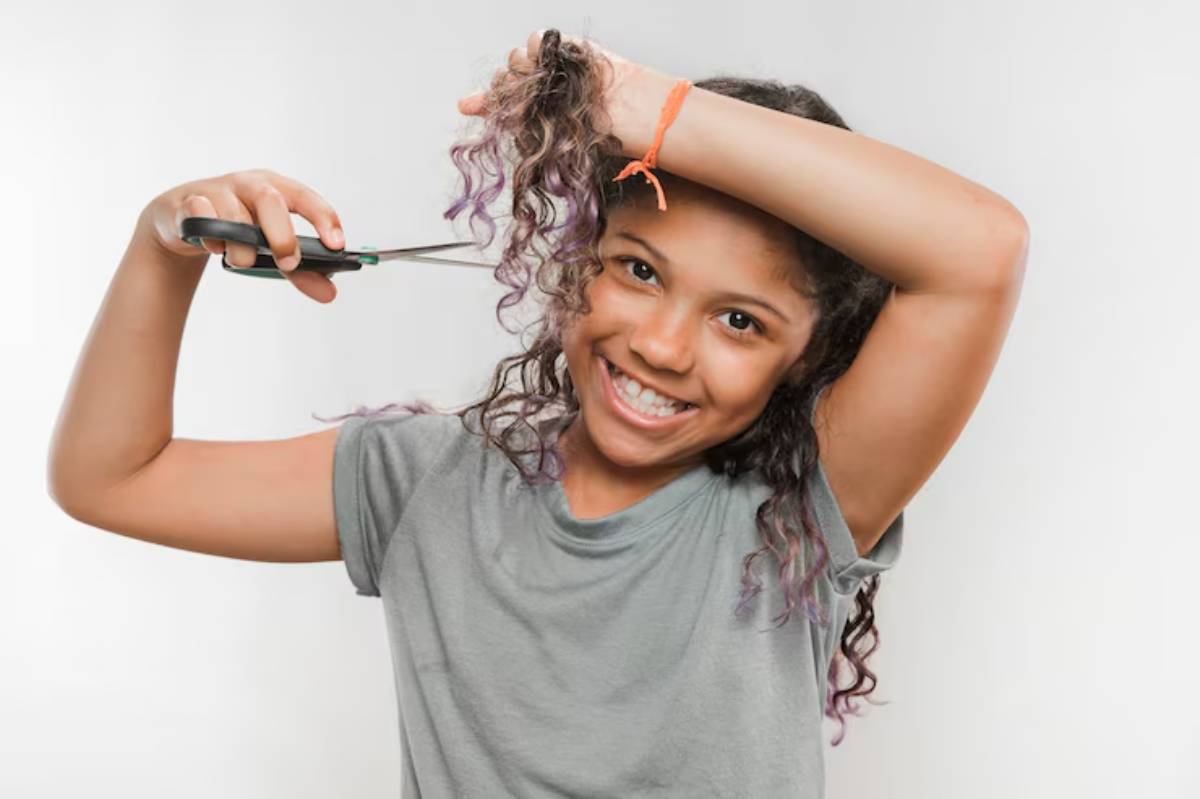 A person with curly hair holds scissors, preparing to cut their hair while wearing a gray t-shirt against a plain background. 