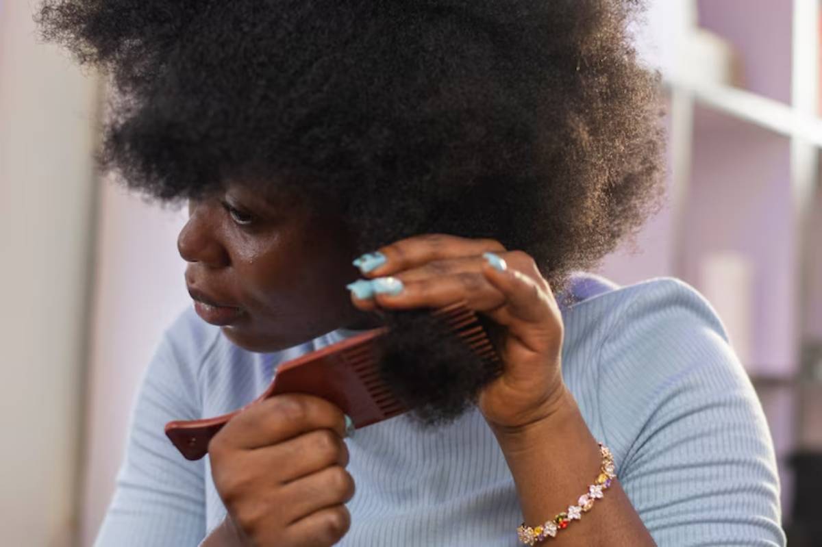 A close-up of a person's hand holding a wooden comb, styling dense, voluminous natural hair against a softly blurred background.