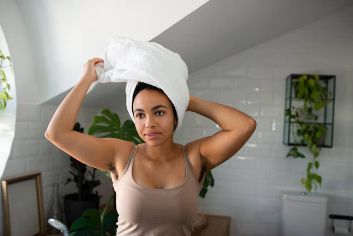 A woman in a cozy bathroom wraps a white towel around her head, with plants and modern decor visible in the background.
