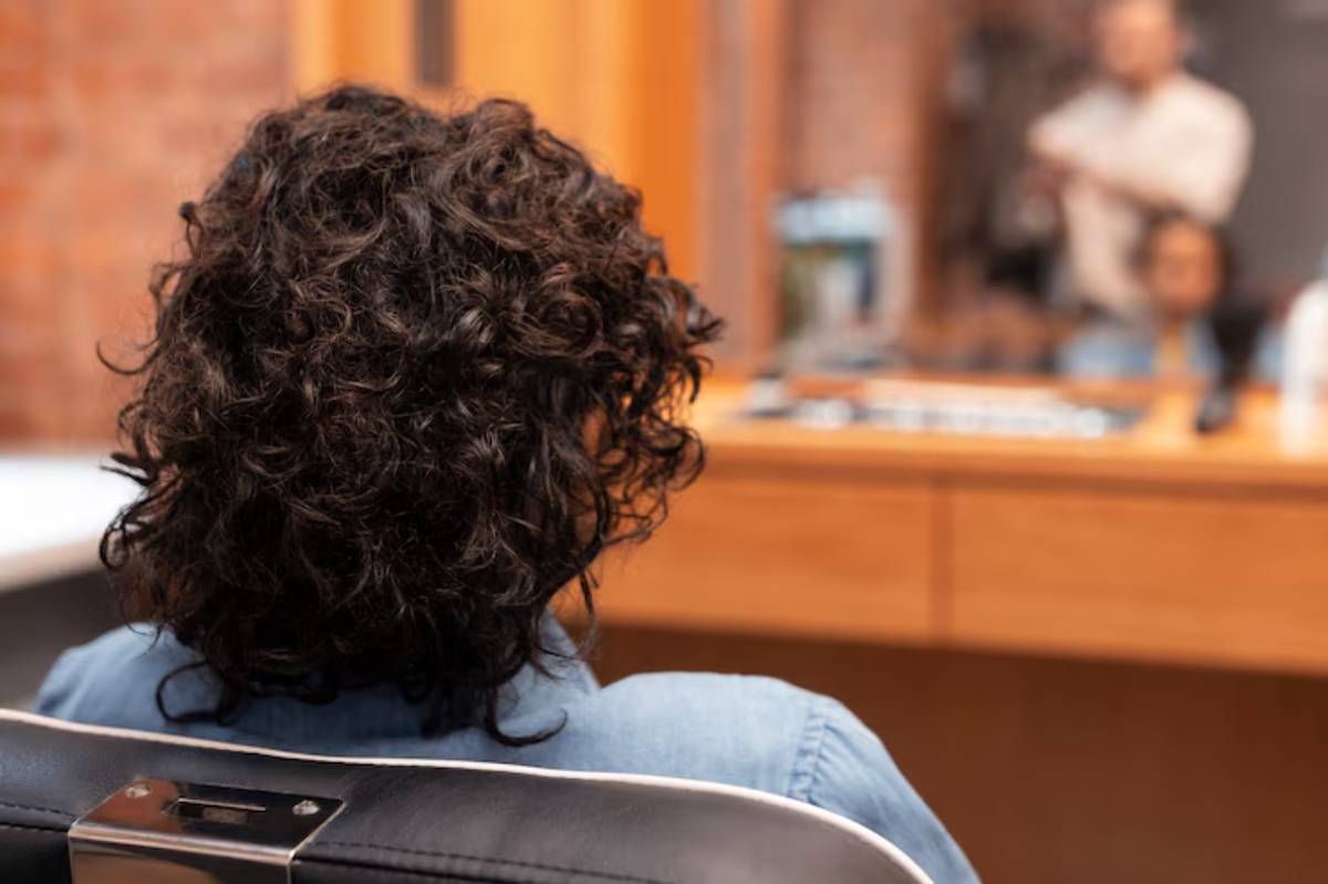 A person with curly hair sits in a salon chair, facing a wooden counter, as a stylist prepares to cut their hair. 