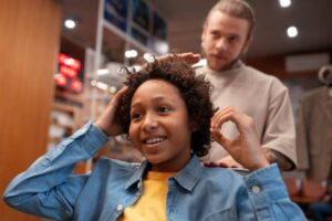 A person with curly hair sits in a salon chair, while a stylist gently works on their hair in a lively barbershop environment.