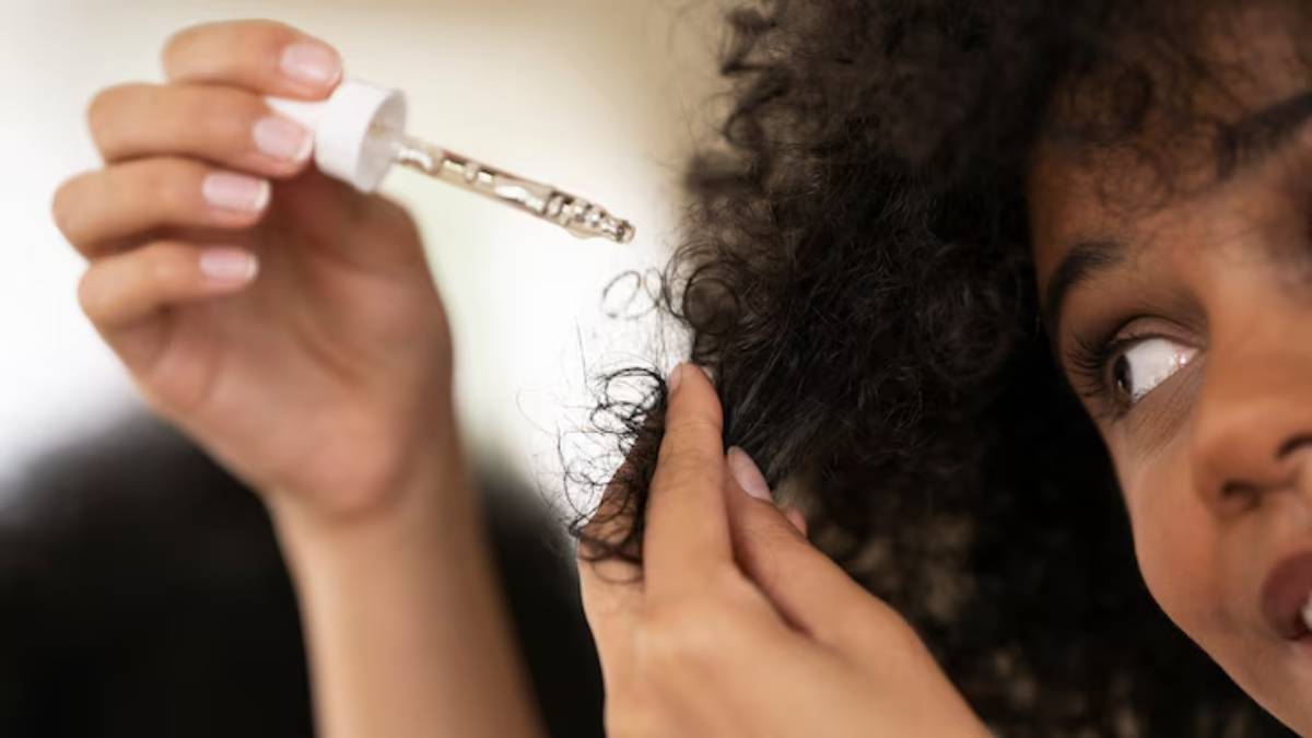 A woman applies a nourishing hair serum from a dropper to her curly hair, focusing intently on the strands. 