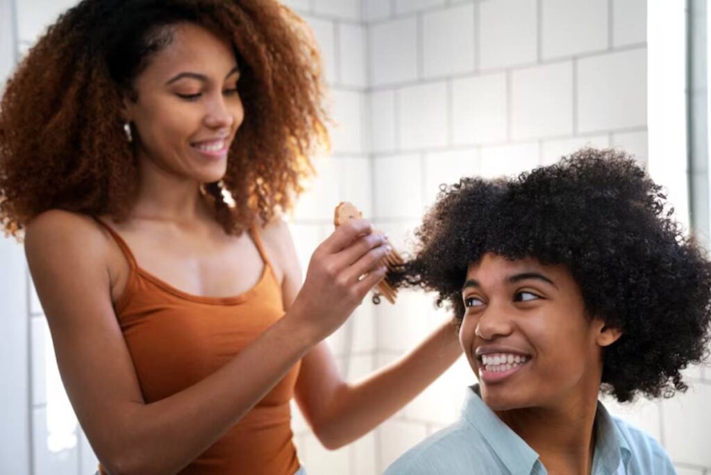 A person with curly hair is being styled by another individual, surrounded by bright, tiled bathroom walls.