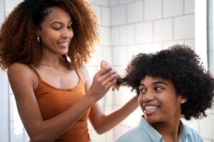 A person with curly hair is being styled by another individual, surrounded by bright, tiled bathroom walls.