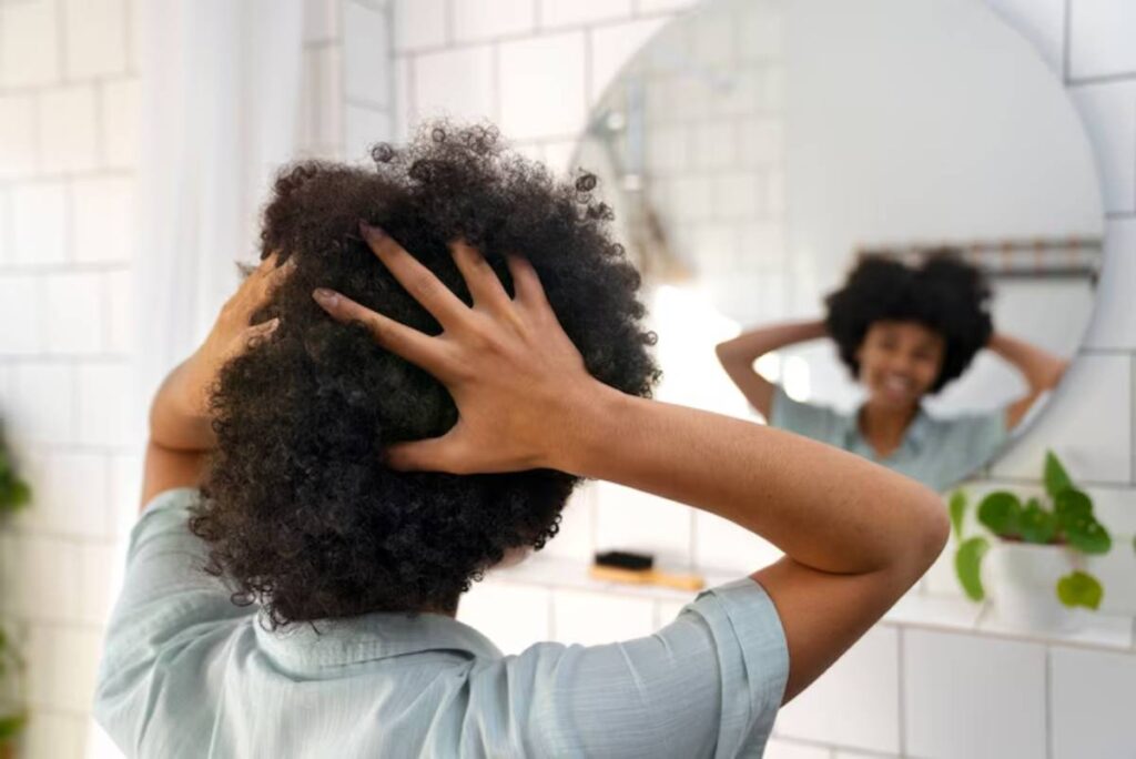 A person with curly hair is shown from behind, holding their head, in front of a circular bathroom mirror. Green plants are visible.