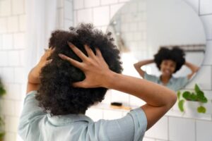 A person with curly hair is shown from behind, holding their head, in front of a circular bathroom mirror. Green plants are visible.